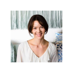 Portrait of a woman with dark hair in a neat, low bun, wearing a white blouse and smiling at the camera. She is seated in front of a decorative background with a woven wall hanging and patterned pillows.