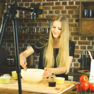 A woman with long blonde hair smiling as she mixes ingredients in a large bowl, set in a rustic kitchen with a brick wall and shelves holding jars and spices. A camera tripod is positioned in the foreground.