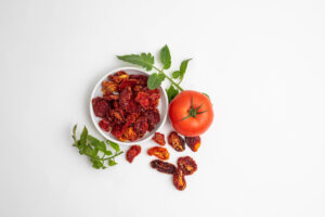 A fresh red tomato and a bowl of dried tomatoes surrounded by green tomato leaves, placed on a clean white background.