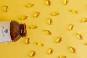 A brown glass bottle labeled 'DM Essentials' tilted on its side, spilling golden capsules onto a bright yellow background.