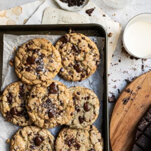 A tray of freshly baked chocolate chip cookies sprinkled with sea salt, surrounded by chocolate chunks, a glass of milk, and vintage recipe papers.