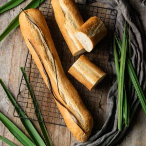 A rustic overhead shot of freshly baked Cuban bread resting on a black cooling rack. The bread has a golden crust with a characteristic deep slit down the center. Surrounding the bread are long green leaves and a dark cloth, creating a moody and artisanal presentation.