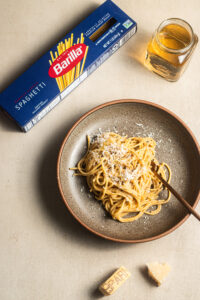 A plate of spaghetti sprinkled with grated Parmesan cheese served in a brown ceramic bowl on a beige surface. A box of Barilla spaghetti and a small jar of olive oil are placed nearby, with chunks of Parmesan cheese scattered around.