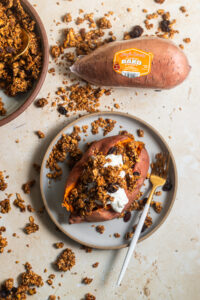 A baked sweet potato topped with whipped cream and granola, served on a light gray plate with a gold and white fork. A whole sweet potato labeled 'Single Sweets Microwaveable Bako Sweet' is placed nearby, surrounded by scattered granola pieces on a light beige surface.