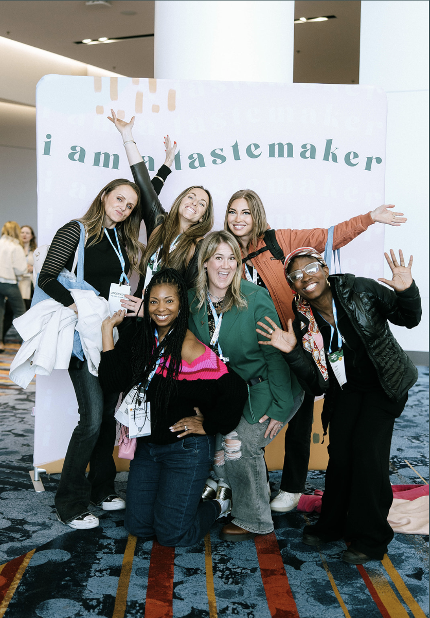 A group of six diverse women pose excitedly in front of a pastel-colored backdrop that reads 'I am a Tastemaker.'