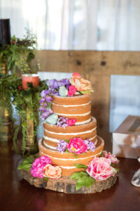 A three-tiered naked wedding cake with layers of golden sponge and thin layers of buttercream frosting. The cake is adorned with vibrant pink, purple, and peach flowers and rests on a wooden slab. It is set on a table with rustic greenery, candles, and a wooden backdrop, creating a warm, natural ambiance.