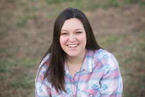 A young woman with long brown hair smiles warmly at the camera. She is wearing a pastel plaid button-up shirt and is sitting outdoors with a soft-focus grassy background. The natural lighting highlights her cheerful expression.