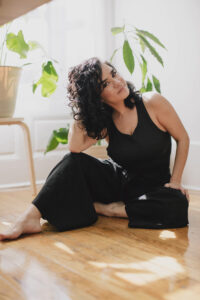 A woman with curly dark hair wearing a black sleeveless top and wide-leg pants, sitting on a wooden floor in a sunlit room surrounded by green houseplants, with a relaxed and thoughtful expression.