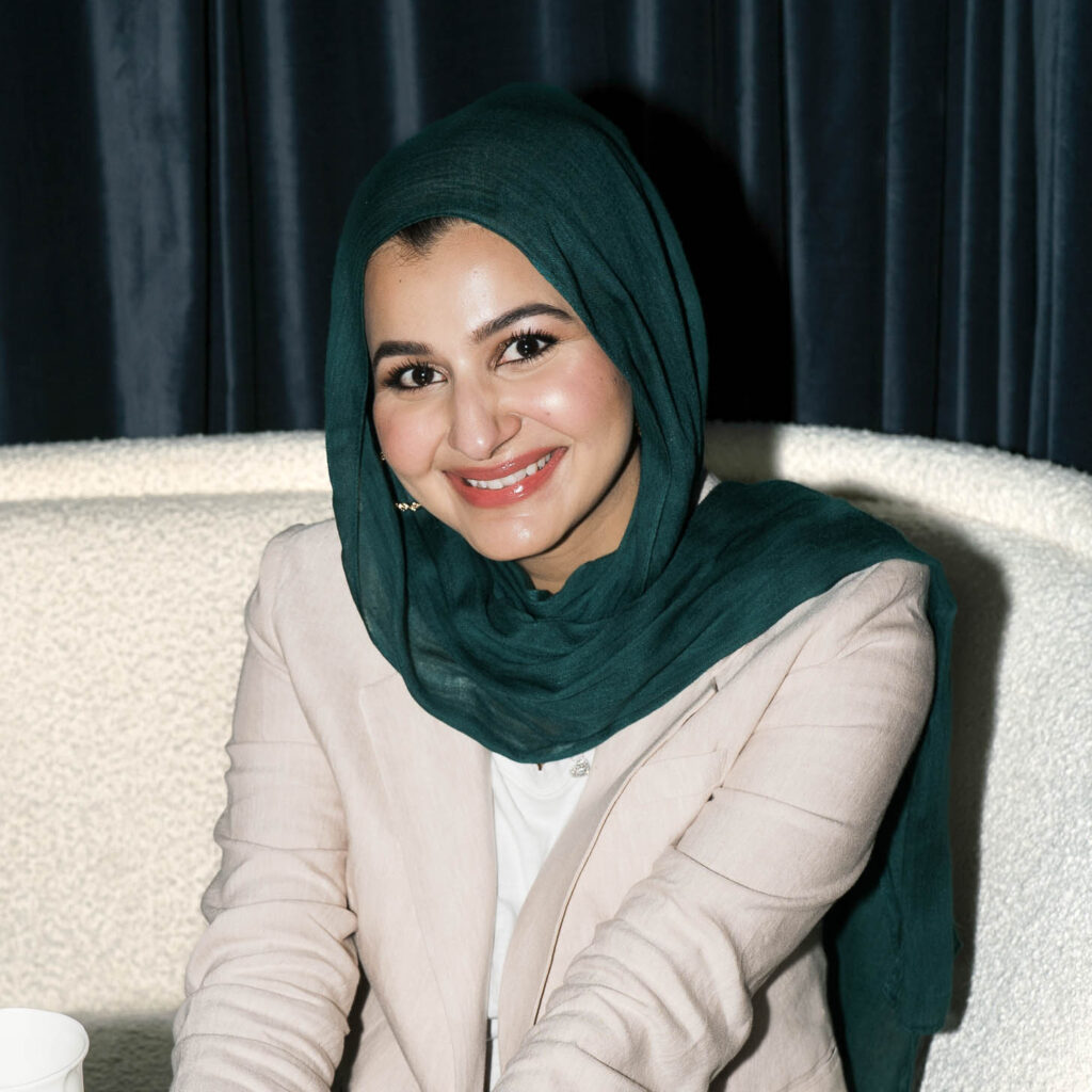 A woman wearing a dark green hijab and a light beige blazer smiles warmly while seated indoors against a textured cream-colored chair and dark curtain backdrop.