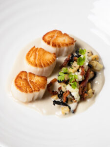 Elegant plated dish featuring three seared scallops with a golden crust, served over a creamy white sauce, accompanied by a garnish of sautéed vegetables and herbs arranged artfully on a white dish.