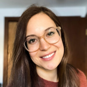 Close-up portrait of a smiling woman with long brown hair, wearing clear-framed glasses and a rust-colored sweater, against a softly lit indoor background.