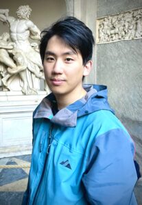 A young man with short black hair wearing a blue and teal outdoor jacket stands in front of a classical marble statue.