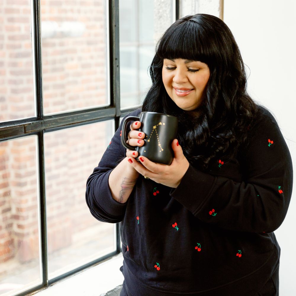 Carla Contreras, a Latina woman with long black hair and bangs, smiles softly while holding a black mug with a gold constellation design. She wears a black sweater with small cherry prints and has red nail polish. She stands by a large industrial-style window with exposed brick in the background, basking in natural light.