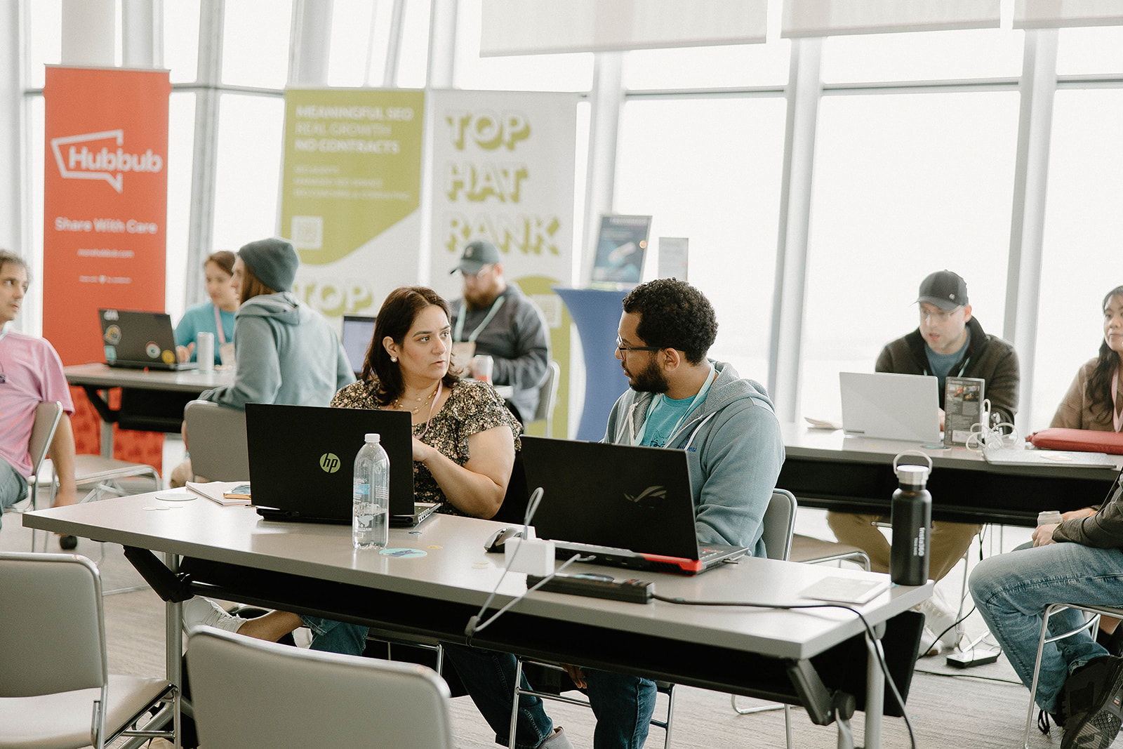 Two attendees talking at a conference table with laptops, surrounded by others working and chatting in a bright, modern space.