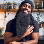 Andrew Bernard smiling warmly, wearing a black beanie and T-shirt, with a long beard, seated at a wooden table in a kitchen with shelves of jars and plants in the background.