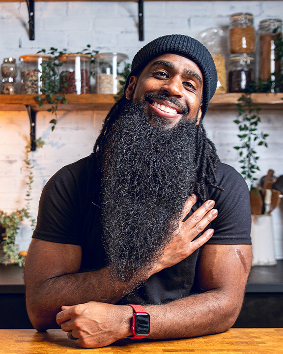 Andrew Bernard smiling warmly, wearing a black beanie and T-shirt, with a long beard, seated at a wooden table in a kitchen with shelves of jars and plants in the background.