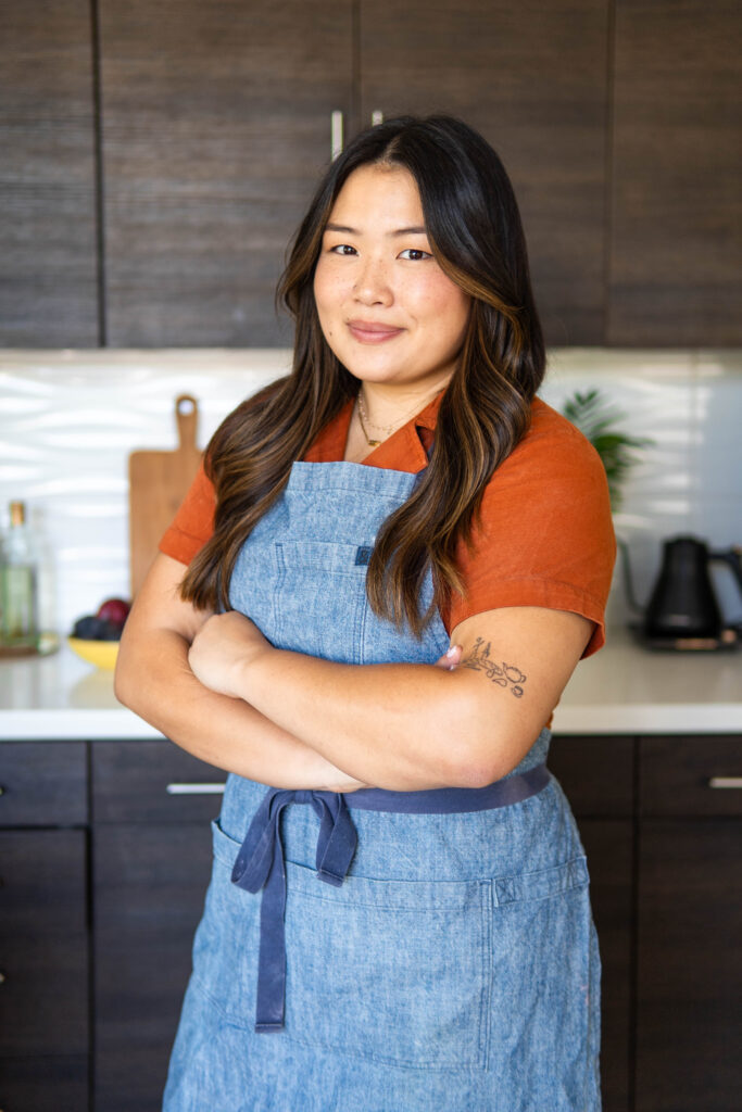 Dara Yu standing confidently in a kitchen, wearing a denim apron over an orange shirt, with arms crossed and a slight smile.