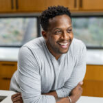 Kevin Curry smiling and leaning on a kitchen counter, wearing a light gray long-sleeve shirt, with wooden cabinets and windows in the background.