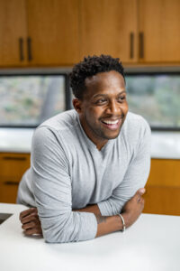Kevin Curry smiling and leaning on a kitchen counter, wearing a light gray long-sleeve shirt, with wooden cabinets and windows in the background.