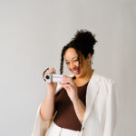 Natasha Pierre smiling while holding a video camera, wearing a white blazer over a brown top, against a neutral background.