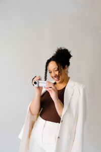 Natasha Pierre smiling while holding a video camera, wearing a white blazer over a brown top, against a neutral background.