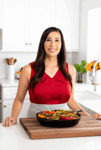 Jessica Gavin smiling with long dark hair wearing a sleeveless red top and light gray apron, standing in a bright white kitchen behind a wooden cutting board with a skillet of colorful stuffed bell peppers.