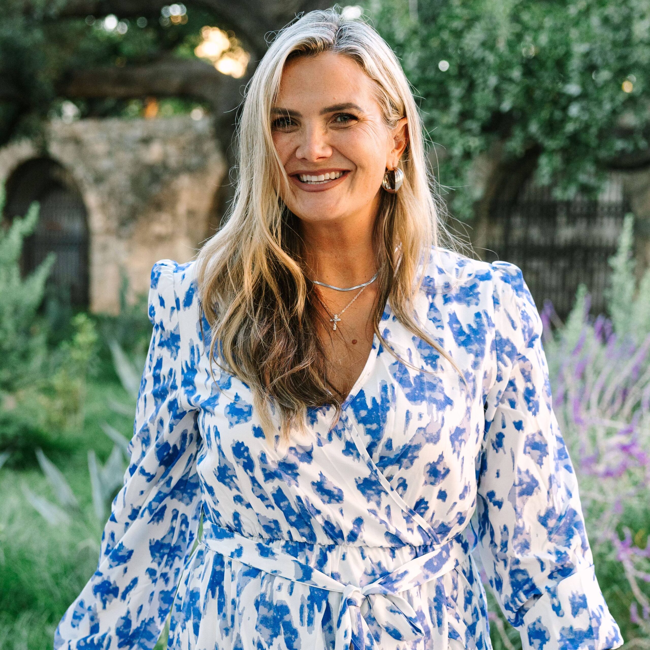 Chandice Probst smiling outdoors while walking through a garden, wearing a blue and white floral dress with greenery and stone arches in the background.