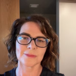Headshot of Heather Wilkins, a woman with short wavy brown hair and black glasses, wearing a black top.