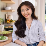 A woman stands in a kitchen smiling. Behind her you can see open-concept shelving with dishware, and there are also dishes on the counter in serving bowls. She wears a button-down shirt and has long dark hair.