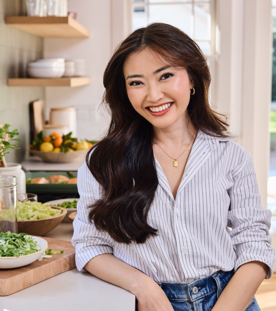 A woman stands in a kitchen smiling. Behind her you can see open-concept shelving with dishware, and there are also dishes on the counter in serving bowls. She wears a button-down shirt and has long dark hair.