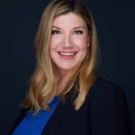 Professional headshot of Jenny Guy smiling, wearing a blue top and black blazer, with long blonde hair styled straight and a dark studio background.
