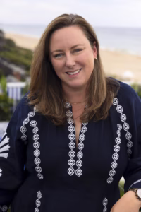 Portrait of Amanda Martin smiling outdoors near the coast, wearing a navy blue embroidered blouse, with the ocean and sandy beach softly blurred in the background.