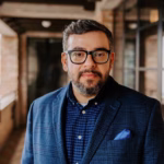 A man with short brown hair, glasses, and a trimmed beard stands in a softly lit hallway. He is wearing a blue plaid blazer, a blue checkered shirt, and a blue pocket square. He looks directly at the camera with a calm, confident expression.
