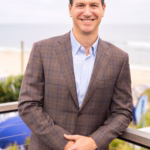 Eric Hochberger smiles at the camera while standing outdoors near the ocean, wearing a brown plaid blazer over a light blue button-down shirt, with his hands resting on a railing and a beachside background softly blurred behind him.