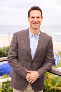Eric Hochberger smiles at the camera while standing outdoors near the ocean, wearing a brown plaid blazer over a light blue button-down shirt, with his hands resting on a railing and a beachside background softly blurred behind him.